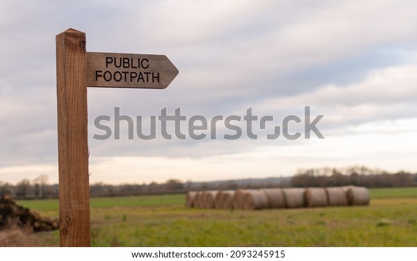 Wooden public footpath sign with\
blurred hay bales in background, UK countryside\
scene
