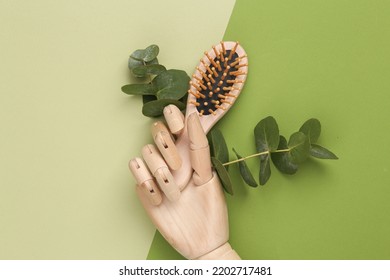 Wooden Puppet Hand Holds Eco Bamboo Hair Brush On A Green Background With A Sprig Of Eucalyptus. Natural Hair Care. Environmentally Friendly