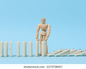 A wooden puppet toy holds back a falling domino on a blue background, representing the concept of a strong personality - Shutterstock ID 2273738457