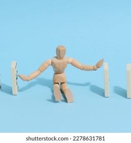 A wooden puppet toy holds back a falling domino on a blue background, representing the concept of a strong personality - Shutterstock ID 2278631781