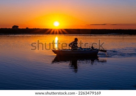 A wooden rowboat against an orange sunset with backlight in the twilight. Silhouette of a 60-year-old Caucasian man with oars in his hands with a poodle dog on a background of sun rays