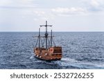 A wooden sailing ship moving through a calm sea under a partly cloudy sky. The ship appears to be an old-fashioned or historical vessel, reminiscent of pirate ships or early exploration vessels. 