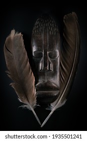 Wooden Sculpture Of Human Face In African Style With Big Feathers Around It. Dark Wood Carving. African Tribal Mask Isolated On Black Background. Art And Culture Of African People. Black History.