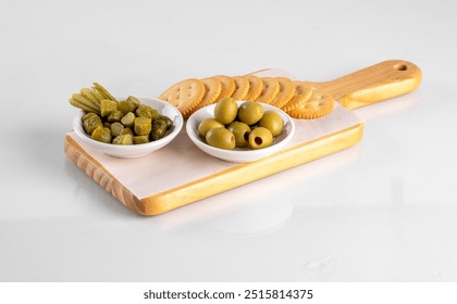 Wooden serving board with green olives, pickles, and crackers on white background. Ideal for appetizers, snacks, and entertaining. - Powered by Shutterstock