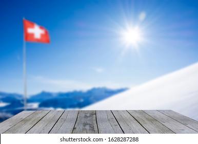 Wooden Shelf Display Table Top Against Snow Covered Blue Mountain Layers With Swiss Flag Fog And Pine Forest On A Sunny Day With Blue Sky