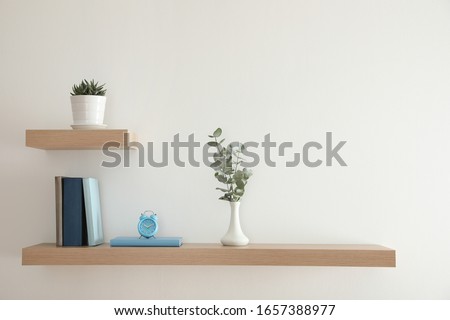 Wooden shelves with beautiful plants, alarm clock and books on light wall