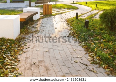 A wooden slatted bench and lamps shaped like large white cubes sit at the edge of a curved walkway in the park.