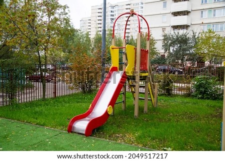 A wooden slide of bright red and yellow color against the background of green trees and residential buildings on a clear sunny day. Playgrounds, sports, entertainment.