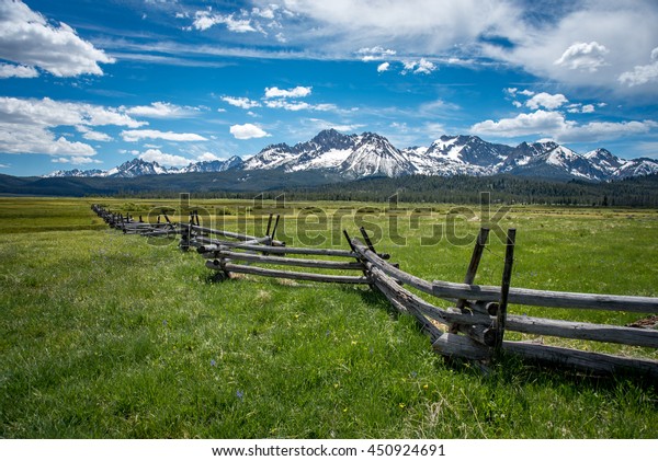 Wooden slit rail fence fades\
into the distance across a green and lush pasture toward the snow\
capped Sawtooth Mountain Range with a blue sky and white clouds\
above.