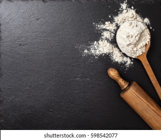  wooden spoon with flour, a rolling pin on a black slate background. Top view, selective focus