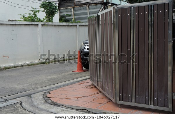 The wooden and stainless steel gate of the house\
is located on the corner of the street. On the road there is a\
traffic cone and a car\
parked.