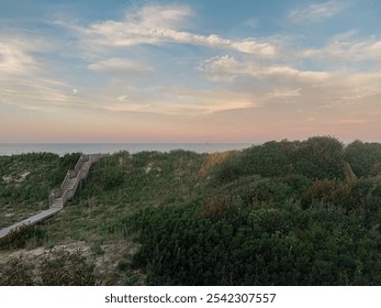 A wooden staircase leads down a grassy dune to a sandy beach at sunset, with a calm ocean and a colorful sky in the background. - Powered by Shutterstock