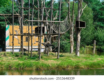 A wooden structure resembling a playground or animal habitat, featuring ropes and nets, surrounded by greenery and a water body. A colorful building is visible in the background. - Powered by Shutterstock