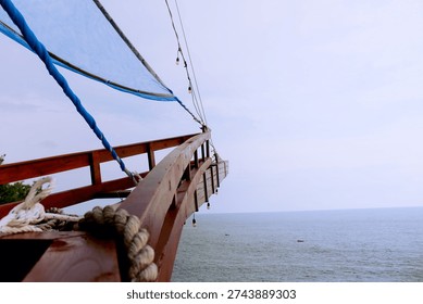 A wooden structure with ropes and string lights stretches toward the cloudy sky, overlooking a calm blue sea. Small boats float in the distance, creating a peaceful coastal scene.