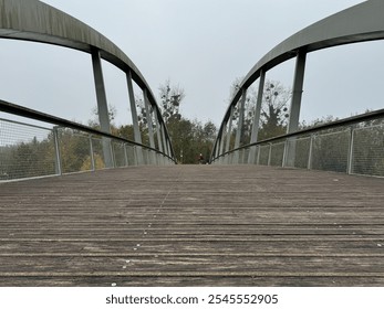 The wooden surface of a pedestrian bridge leads gently into a natural environment, framed by trees under a gray sky. A person can be seen in the distance, enjoying the serene pathway. - Powered by Shutterstock