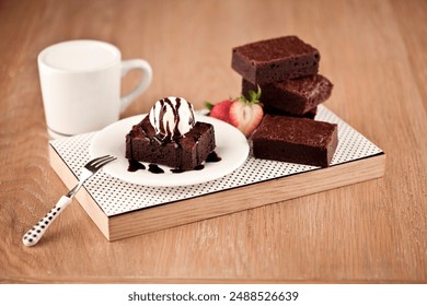 wooden surface, a white plate with black polka dots holds a brownie topped with vanilla ice cream and chocolate syrup. Three more brownies stack beside it, and a halved strawberry shows its texture. - Powered by Shutterstock
