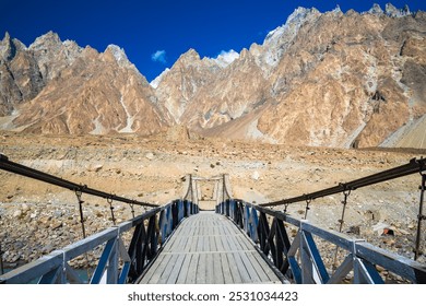 A wooden suspension bridge leads to a majestic backdrop of jagged, snow-capped mountains under a clear blue sky, showcasing rugged natural beauty and adventure in the wilderness. - Powered by Shutterstock