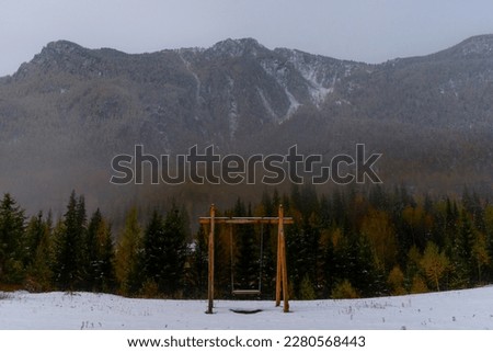A wooden swing by the forest on the snow under a mountain with fog and white snow, with the light of the moon on a bright night in Altai in Siberia.