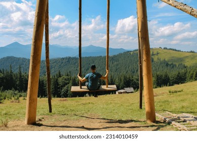 Wooden Swing on Mountain Meadow: A Serene Retreat with Majestic Mountain View and Lush Pine Forest. Enjoy a Sunny Summer Day as a man Delights in Swinging After a Rewarding Hike in the Carpathian
