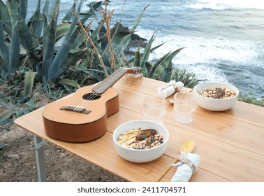 Wooden table with acoustic guitar bowls of muesli cups spoons and folded towels set near green coastal plant on seashore against waving sea - Powered by Shutterstock