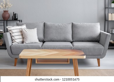 Wooden table and big grey couch with pillows in living room of trendy apartment, real photo with copy space on the wall