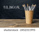 A wooden table with a brown cup and a bunch of pencils. The cup has the word bilingual written on it