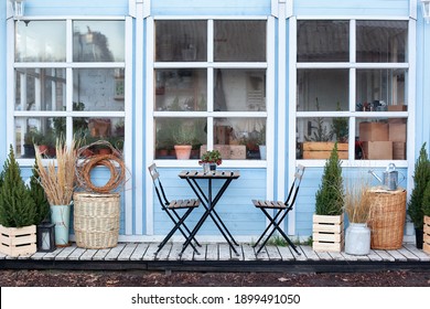 Wooden Table And Chairs On Veranda Of House. Exterior Street Cafe With Furnuture. Outdoor Home Decor. Facade Terrace With Wicker Baskets And Green Plants. Porch Home With Christmas Trees In Pots
