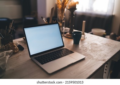 Wooden Table With Cup, Opened Laptop Computer And Many Art Items And Equipment In Dark Art Studio With Sunny Lighting. Working Place Of Artist, Sculptor Or Other Art Worker. No People