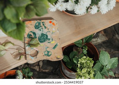 A wooden table displays a color-filled paint palette surrounded by vibrant potted plants. This cozy garden workspace invites creativity and inspiration during a sunny afternoon. - Powered by Shutterstock