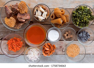 A Wooden Table Filled With Many Small Bowls Of African Food And Nigerian Soup Ingredients Including, Peppers, Vegetables, Dried Fish, Meat, Salt, Oil, Grounded Spices And Seasoning Cubes