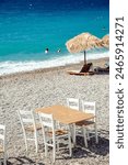 Wooden table with four wooden chairs, on the pebbled beach of Loutraki, Greece, where swimmers enjoy the azure waters during the summer vacation.