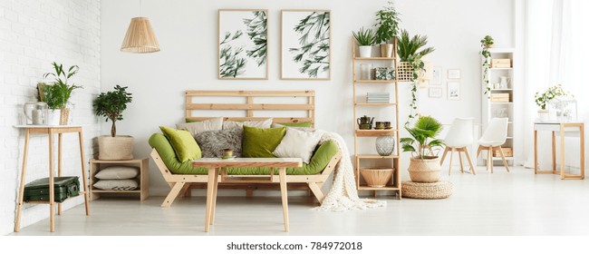 Wooden Table In Front Of Green Couch With Cushions In Floral Living Room Interior With Leaves Posters And Suitcase On Shelf