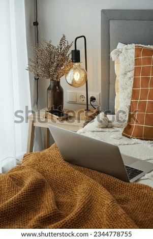 A wooden table with a lamp turned on and decor by the window and a bed with a brown plaid and a laptop