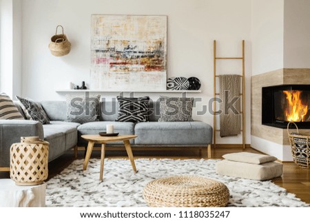 Wooden table next to grey corner settee in warm living room interior with painting and fireplace. Real photo
