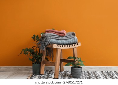 Wooden table with stack of soft blankets and houseplants near orange wall