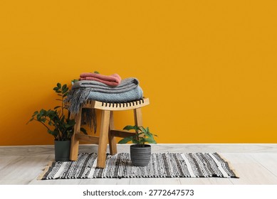 Wooden table with stack of soft blankets and houseplants near orange wall