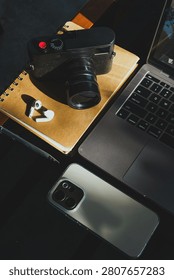 wooden table workspace setup featuring a laptop computer, a plastic cup of iced black coffee, modern smartphone, wireless earbuds, notebook, a pen, classic black rangefinder camera morning sunlight.