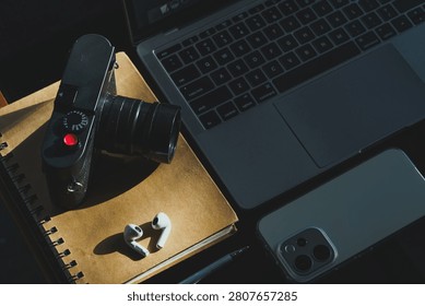 wooden table workspace setup featuring a laptop computer, a plastic cup of iced black coffee, modern smartphone, wireless earbuds, notebook, a pen, classic black rangefinder camera morning sunlight.