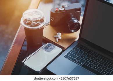 wooden table workspace setup featuring a laptop computer, a plastic cup of iced black coffee, modern smartphone, wireless earbuds, notebook, a pen, classic black rangefinder camera morning sunlight.