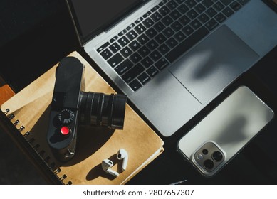 wooden table workspace setup featuring a laptop computer, a plastic cup of iced black coffee, modern smartphone, wireless earbuds, notebook, a pen, classic black rangefinder camera morning sunlight.