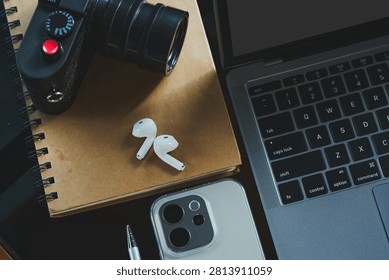 wooden table workspace setup featuring a laptop computer, a plastic cup of iced black coffee, modern smartphone, wireless earbuds, notebook, a pen, classic black rangefinder camera morning sunlight.