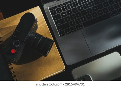 wooden table workspace setup featuring a laptop computer, a plastic cup of iced black coffee, modern smartphone, notebook, a pen, classic black rangefinder camera morning sunlight.