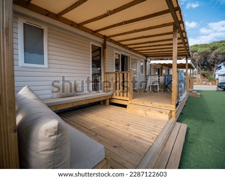 Wooden terrace of a bungalow in a southern France camping with beige pillows, grass, pines trees and cloudy blue sky