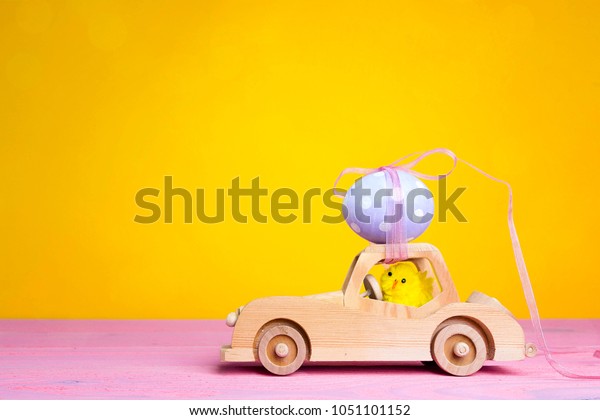Wooden toy car with Easter egg on\
the roof and chick driver on yellow background. Copy\
space.