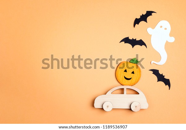 Wooden toy car with funny pumpkin on the roof\
and bats on orange background. Space for text. Flat lay Halloween\
background.