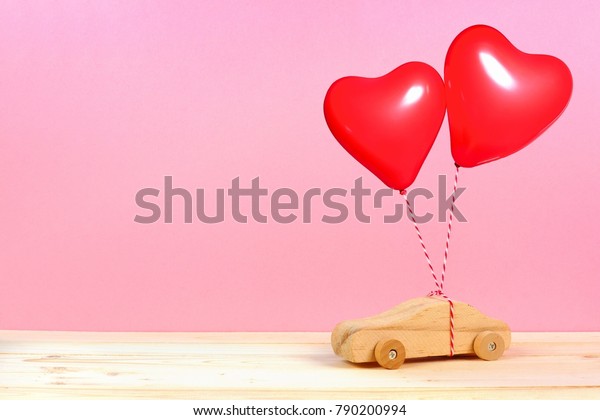 Wooden toy car with red\
heart balloons against a pink background. Valentines Day or love\
concept.