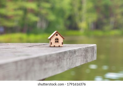 A Wooden Toy House With A Heart Above The Window Stands On A Wooden Table On The Lake Shore . The Concept Of Selling, Buying, Renting Real Estate, Family Home, Home For Children, 