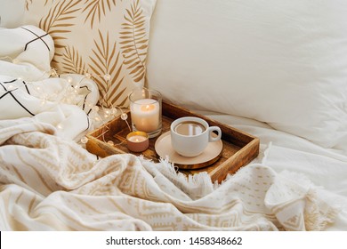 Wooden tray of coffee and candles on bed. White bedding sheets with striped blanket and pillow. Breakfast in bed. Hygge concept.