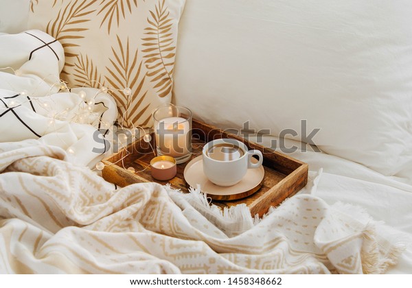 Wooden tray of coffee and candles on bed. White\
bedding sheets with striped blanket and pillow. Breakfast in bed.\
Hygge concept.