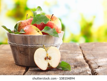 Wooden tub full of freshly harvested red apples with a halved apple on display on a wooden tabletop outdoors with copyspace